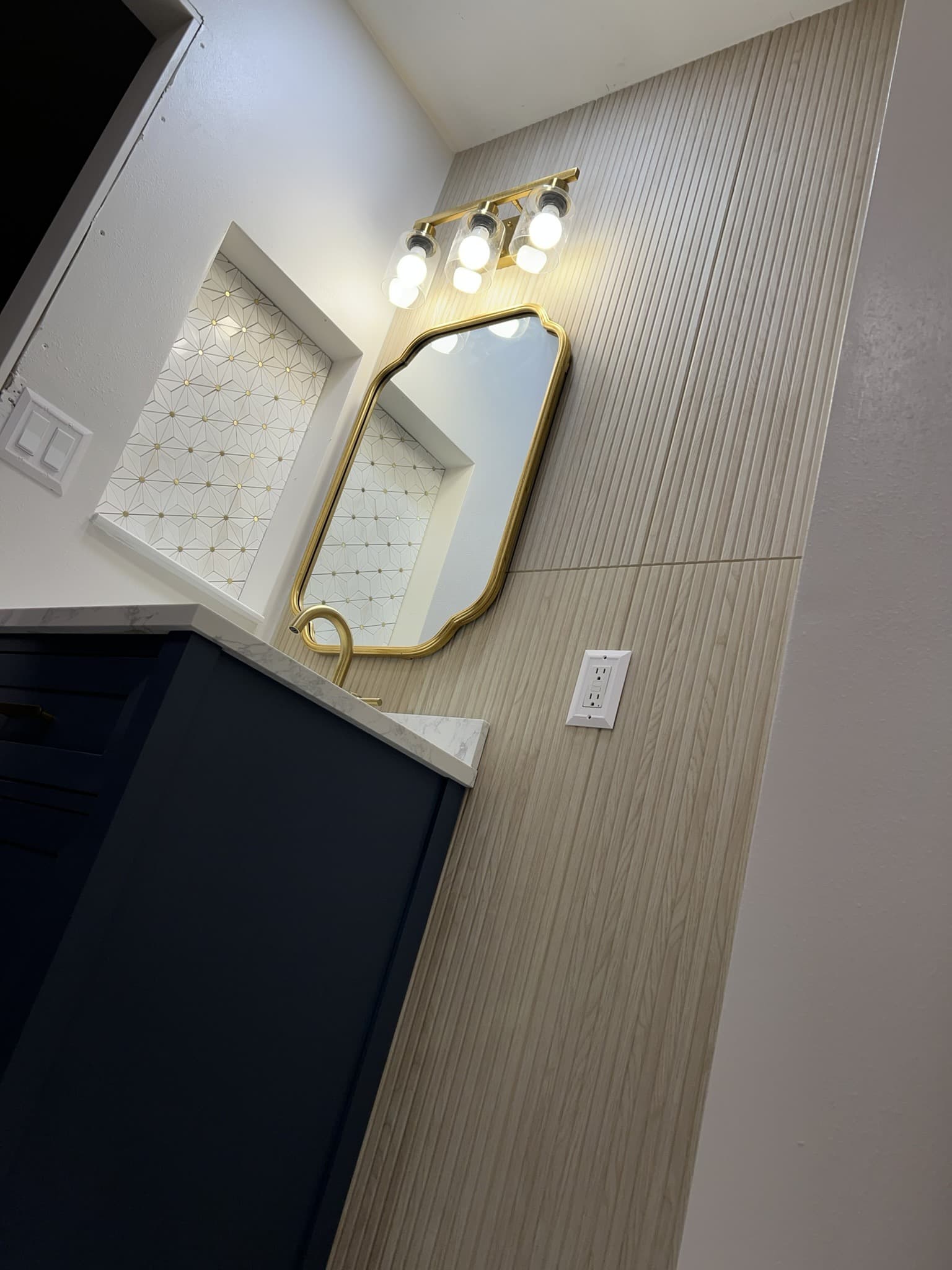 Navy vanity with marble top and arched gold mirror beside a recessed gold-star mosaic niche — Karhan Construction, Orlando