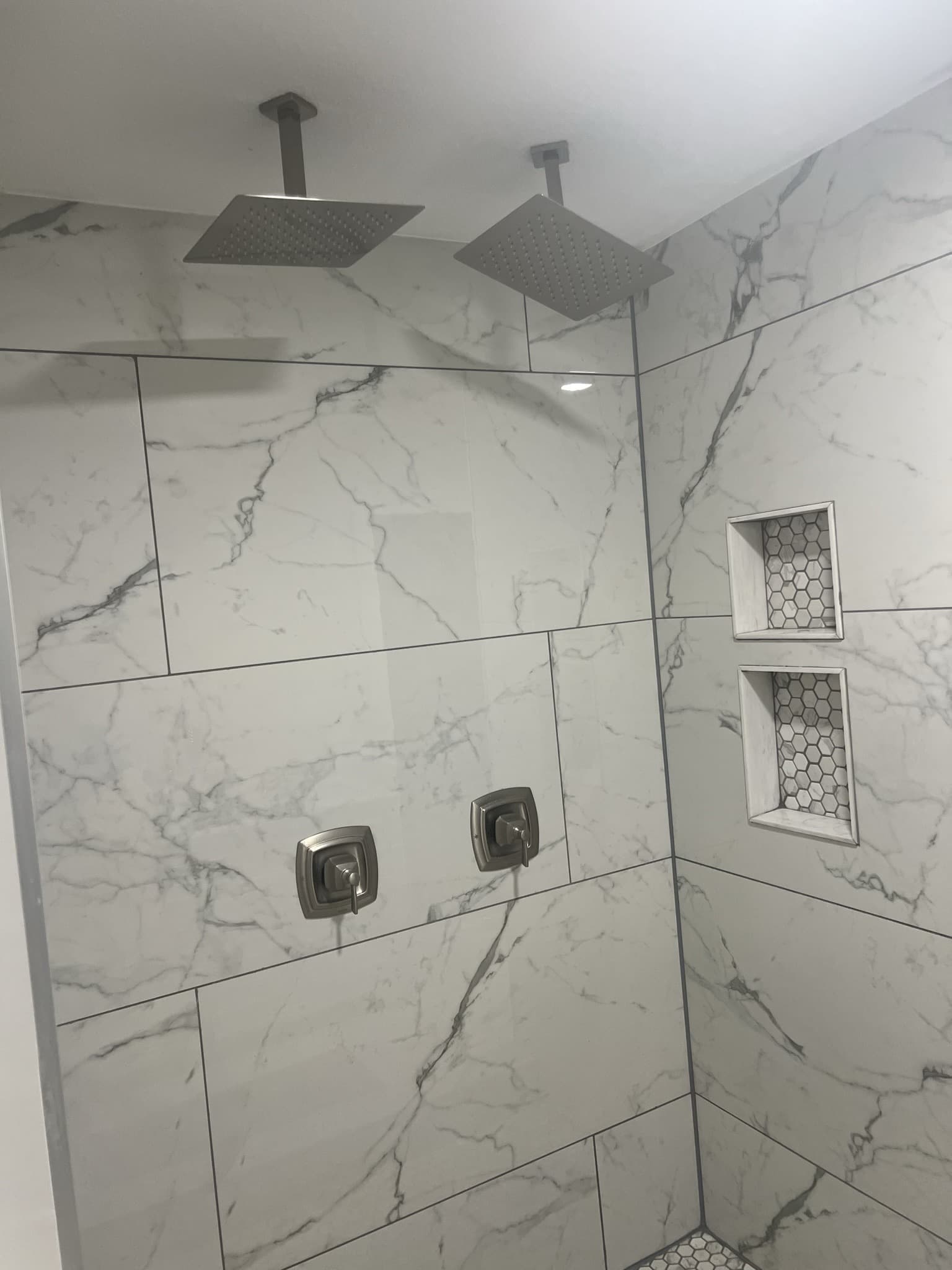 Two ceiling-mounted square rain shower heads on marble-look walls with twin hexagon-mosaic niches — Karhan Construction, Orlando