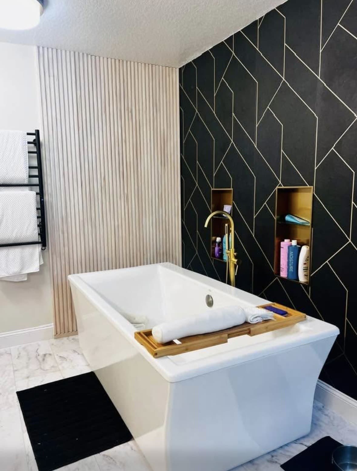Freestanding white soaking tub with bamboo tray, gold floor faucet, and black-and-gold geometric tile wall — Karhan Construction, Orlando