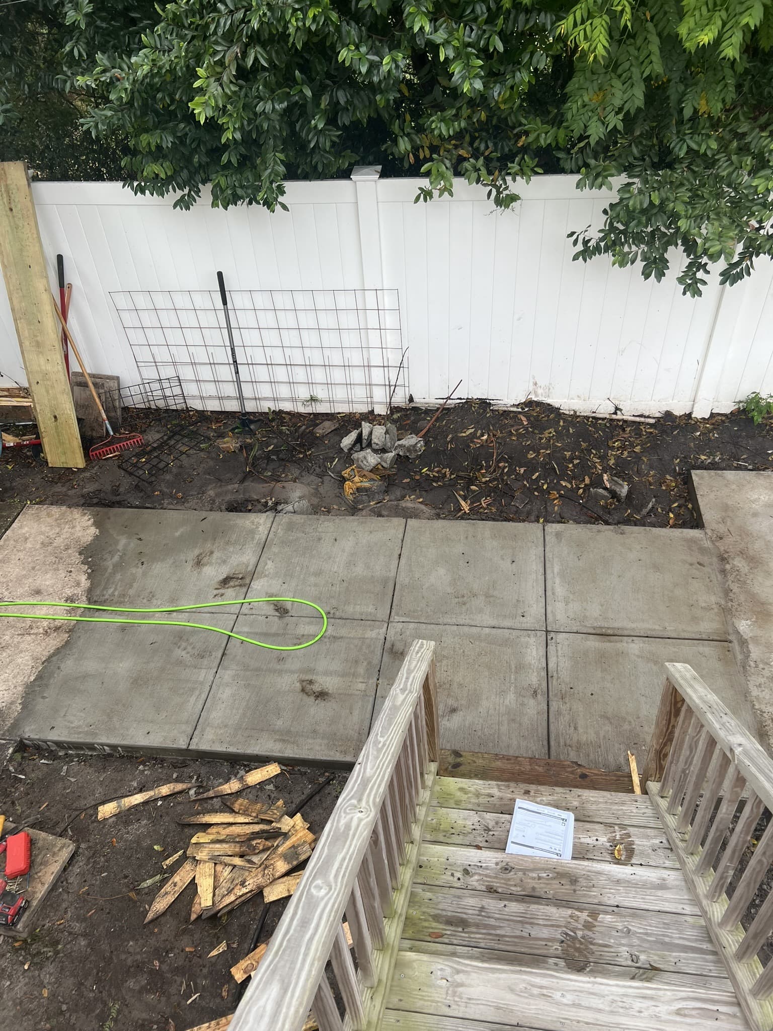 Cured concrete patio pad with tooled control joints viewed from deck steps, garden bed along the white fence — Karhan Construction, Orlando