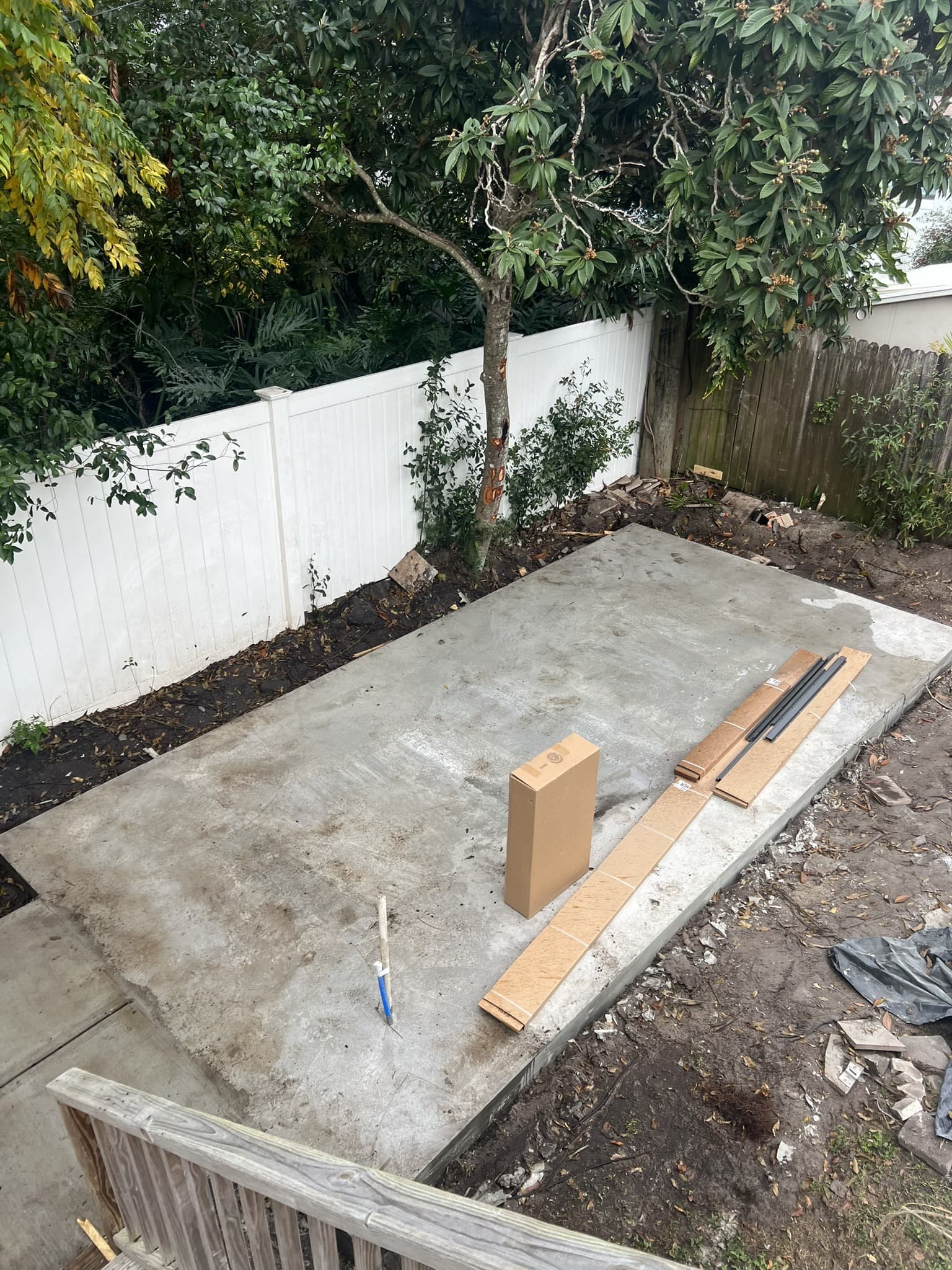 Finished cured concrete slab pad in a fenced backyard beneath shade trees, deck railing in the foreground — Karhan Construction, Orlando