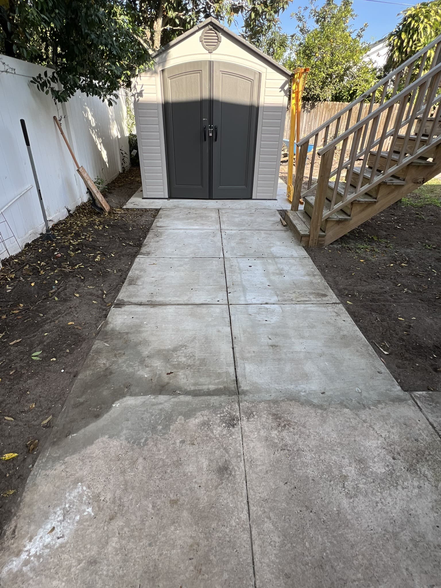 Finished concrete walkway leading to a tan resin storage shed with dark doors beside wooden deck stairs — Karhan Construction, Orlando