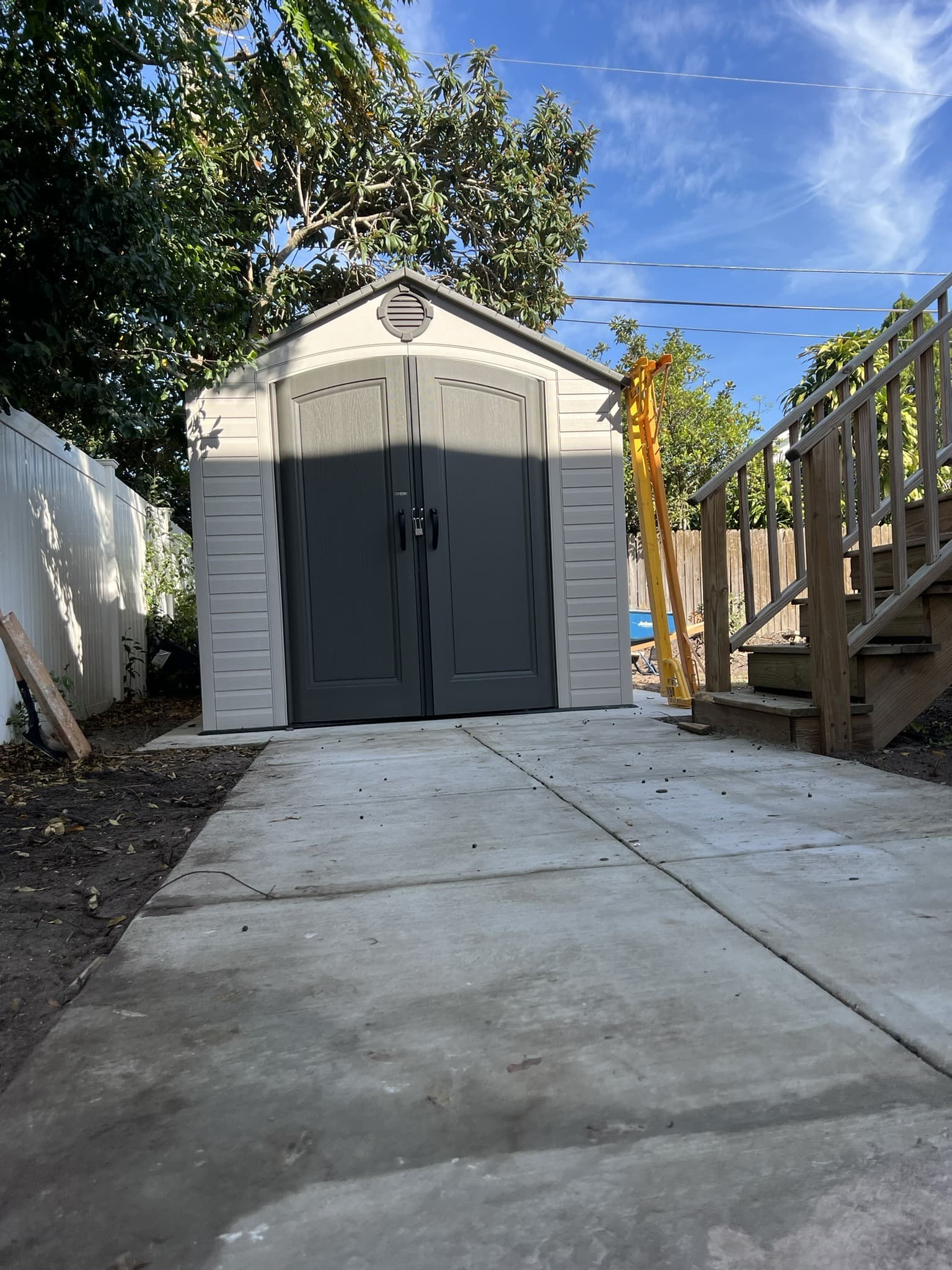 Wide finished concrete pad and walkway leading to a gray resin shed under a blue sky, deck stairs at right — Karhan Construction, Orlando