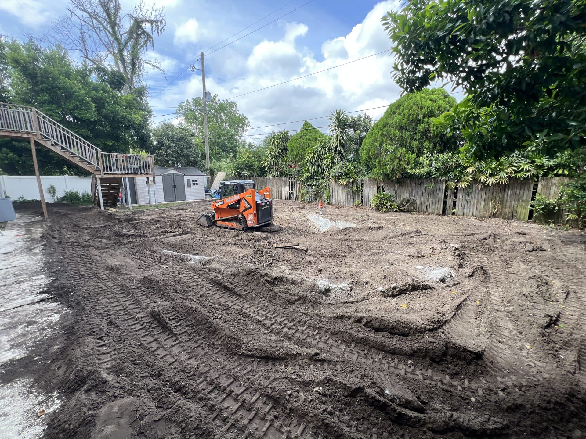Backyard graded to bare dirt with an orange compact track loader parked mid-yard, wood deck stairs at left — Karhan Construction, Orlando