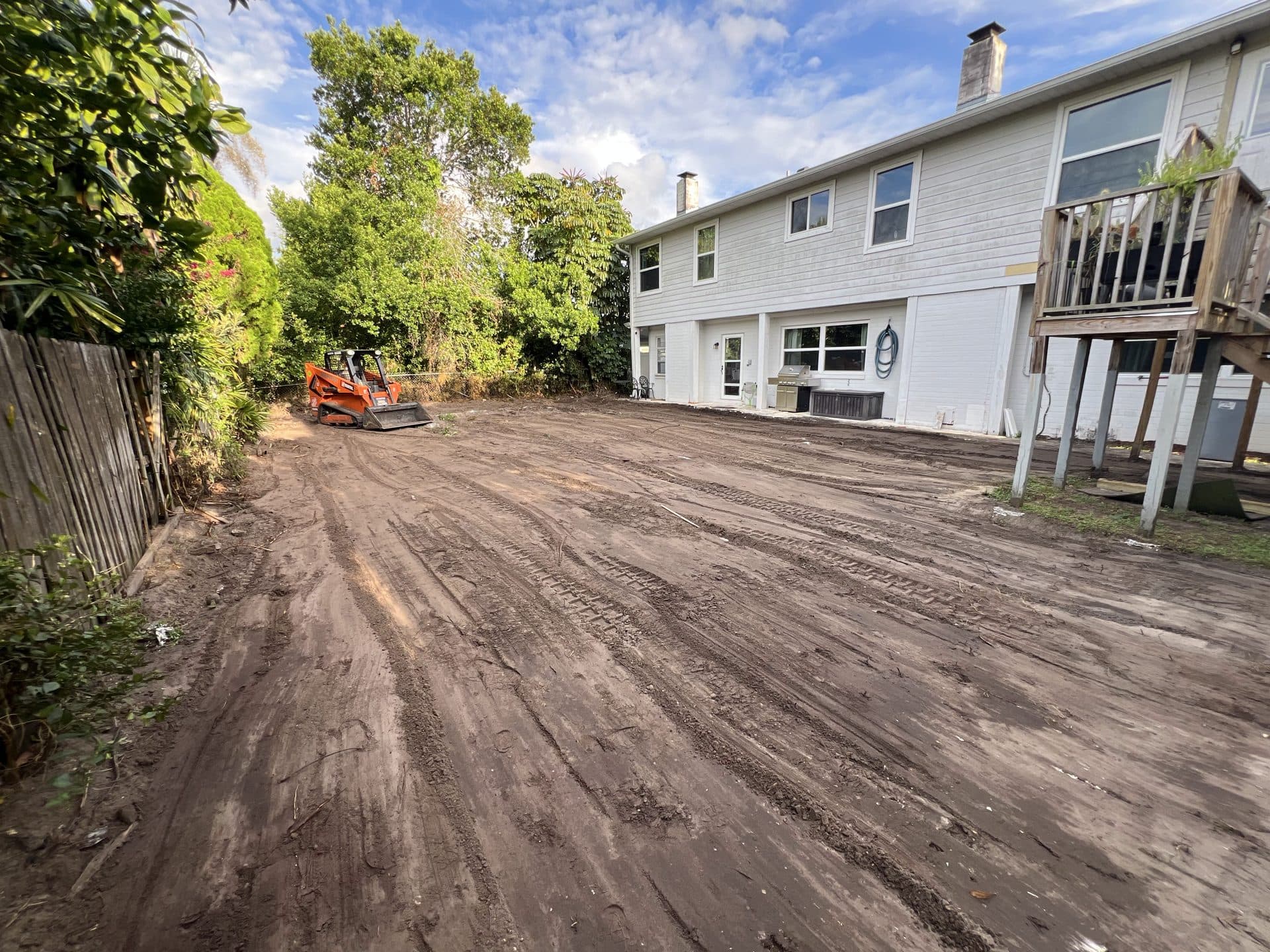 Graded dirt lot behind a large white two-story house, orange compact track loader parked by the tree line — Karhan Construction, Orlando