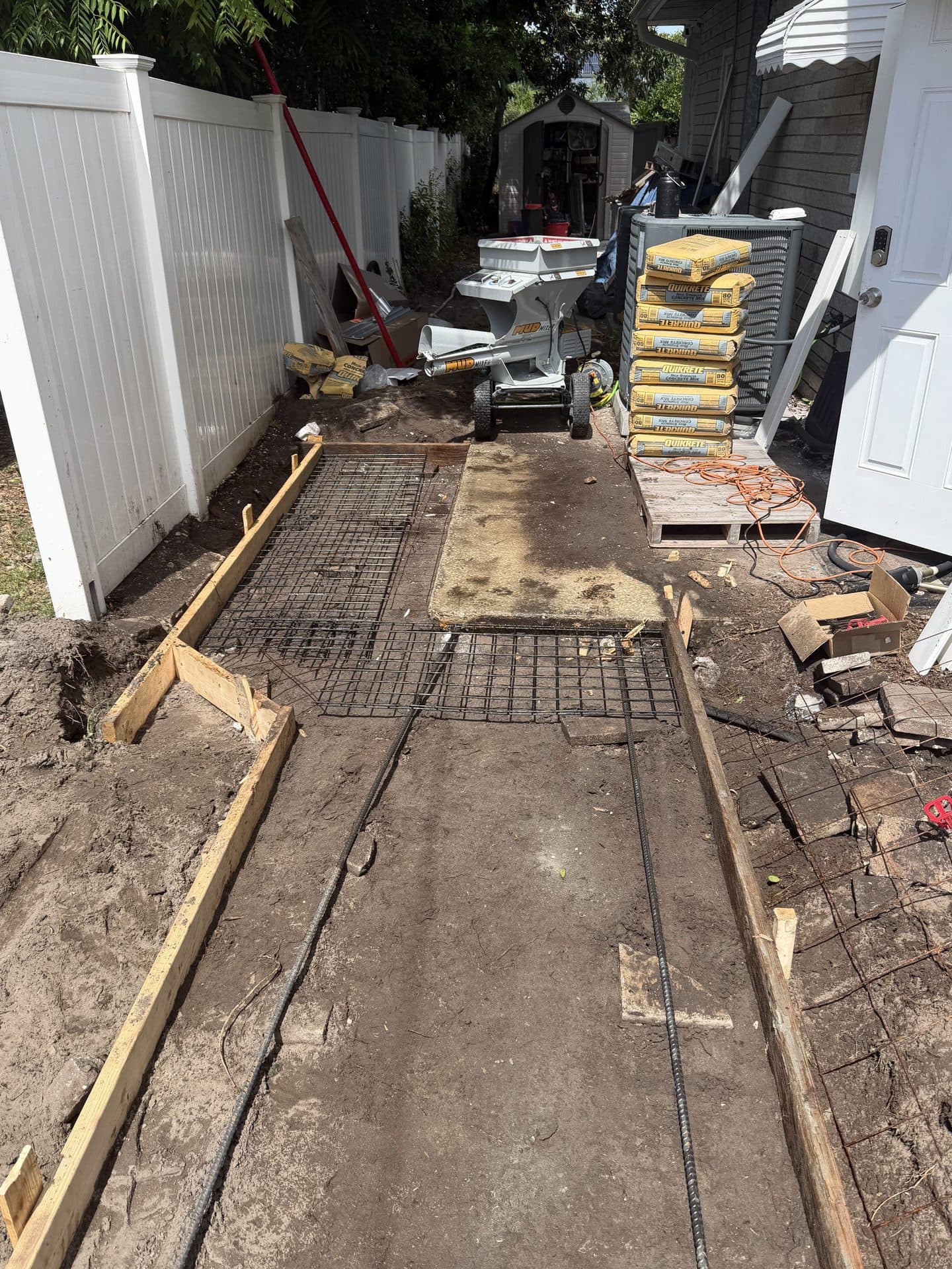 Side-yard walkway forms and wire mesh in soil, stacked Quikrete bags and a mixer by a white vinyl fence — Karhan Construction, Orlando