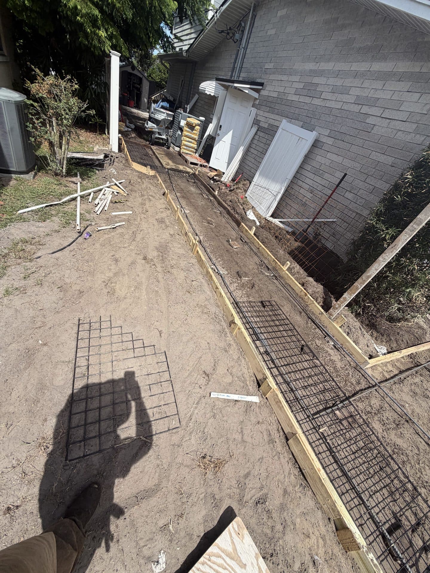 Formed footing trench with rebar and wire mesh dug along a gray block house wall, doors set aside — Karhan Construction, Orlando