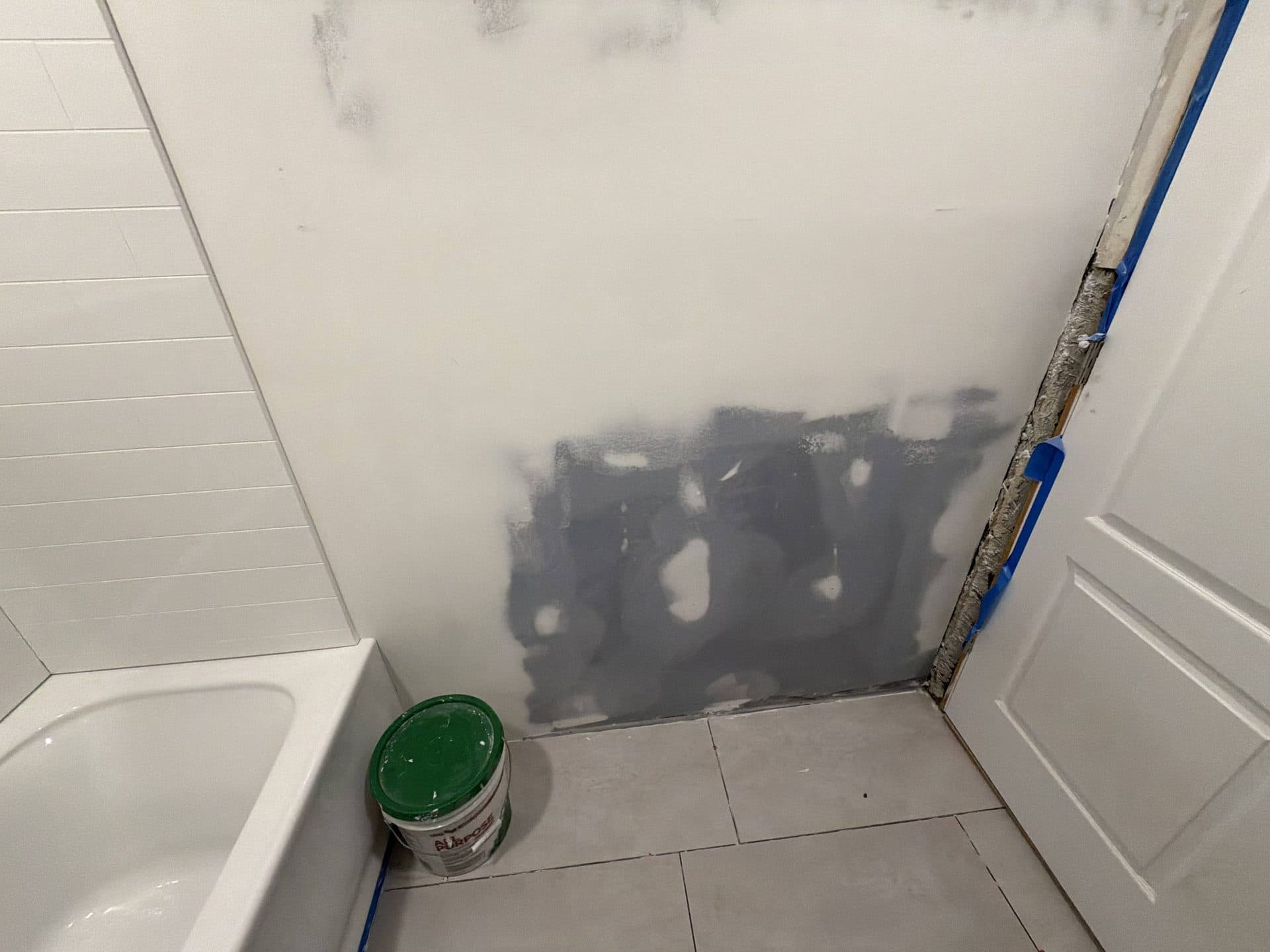 Gray joint compound patched over bathroom drywall beside a white tub and subway tile, bucket on the floor — Karhan Construction, Orlando