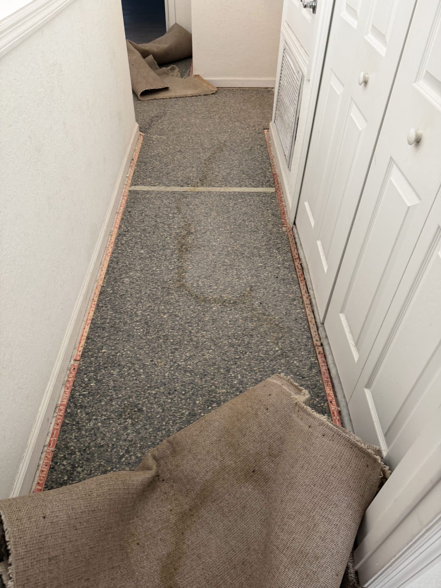 Hallway carpet torn out to expose the gray concrete slab and red tack strips, white louvered doors at right — Karhan Construction, Orlando