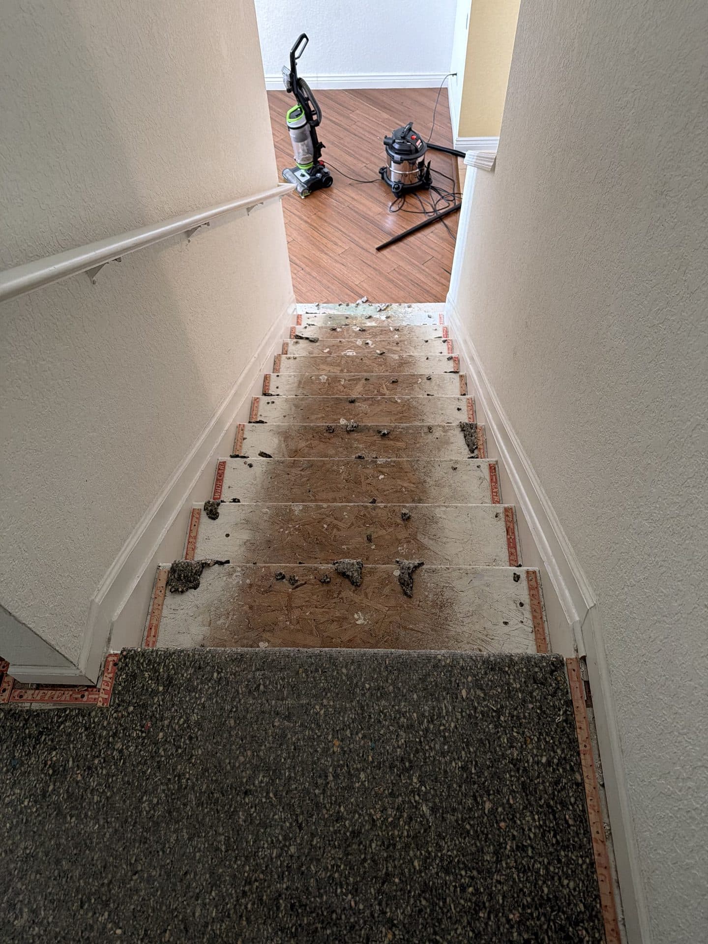 Staircase stripped to bare OSB treads with padding debris and staples, an upright vacuum and shop vac below — Karhan Construction, Orlando