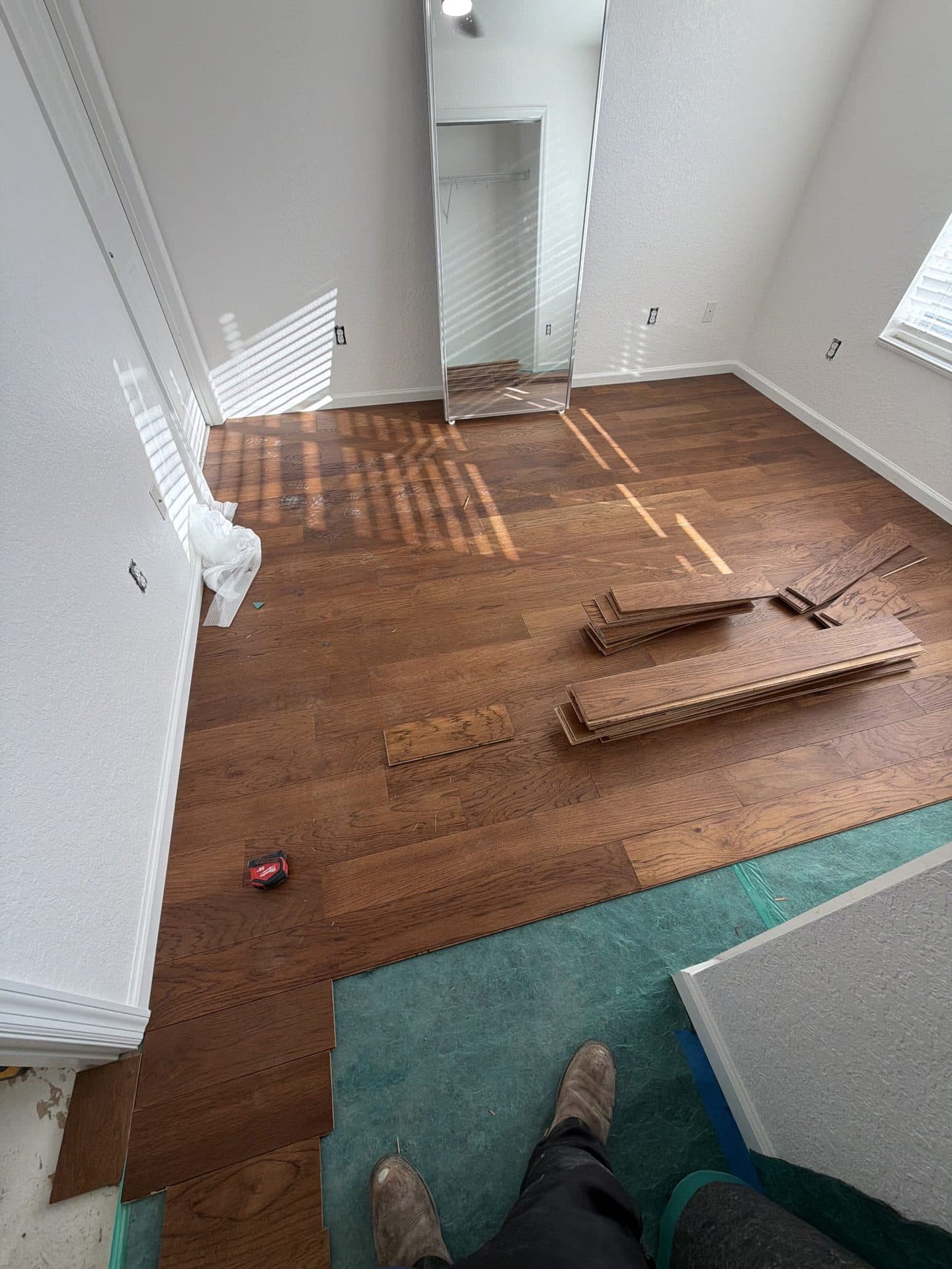Bedroom floor mid-install, medium-brown wood planks laid over green underlayment with loose boards stacked nearby — Karhan Construction, Orlando