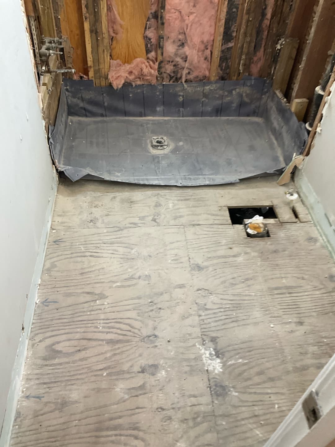 Black shower pan liner set over plywood subfloor with drain flange, studs and pink insulation behind — Karhan Construction, Orlando