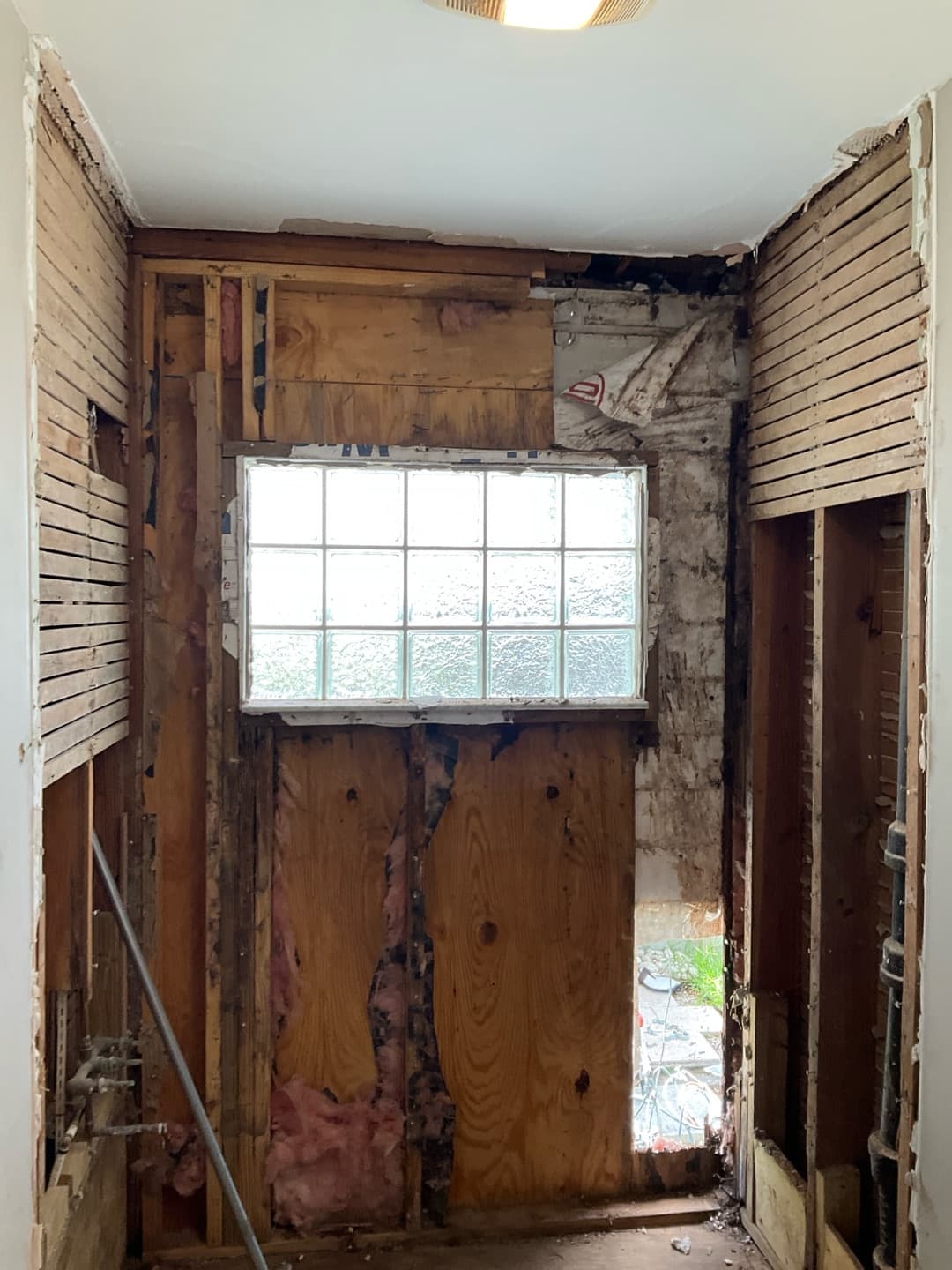 Gutted bathroom showing stud framing and plank sheathing around a glass-block window with a wall opening — Karhan Construction, Orlando
