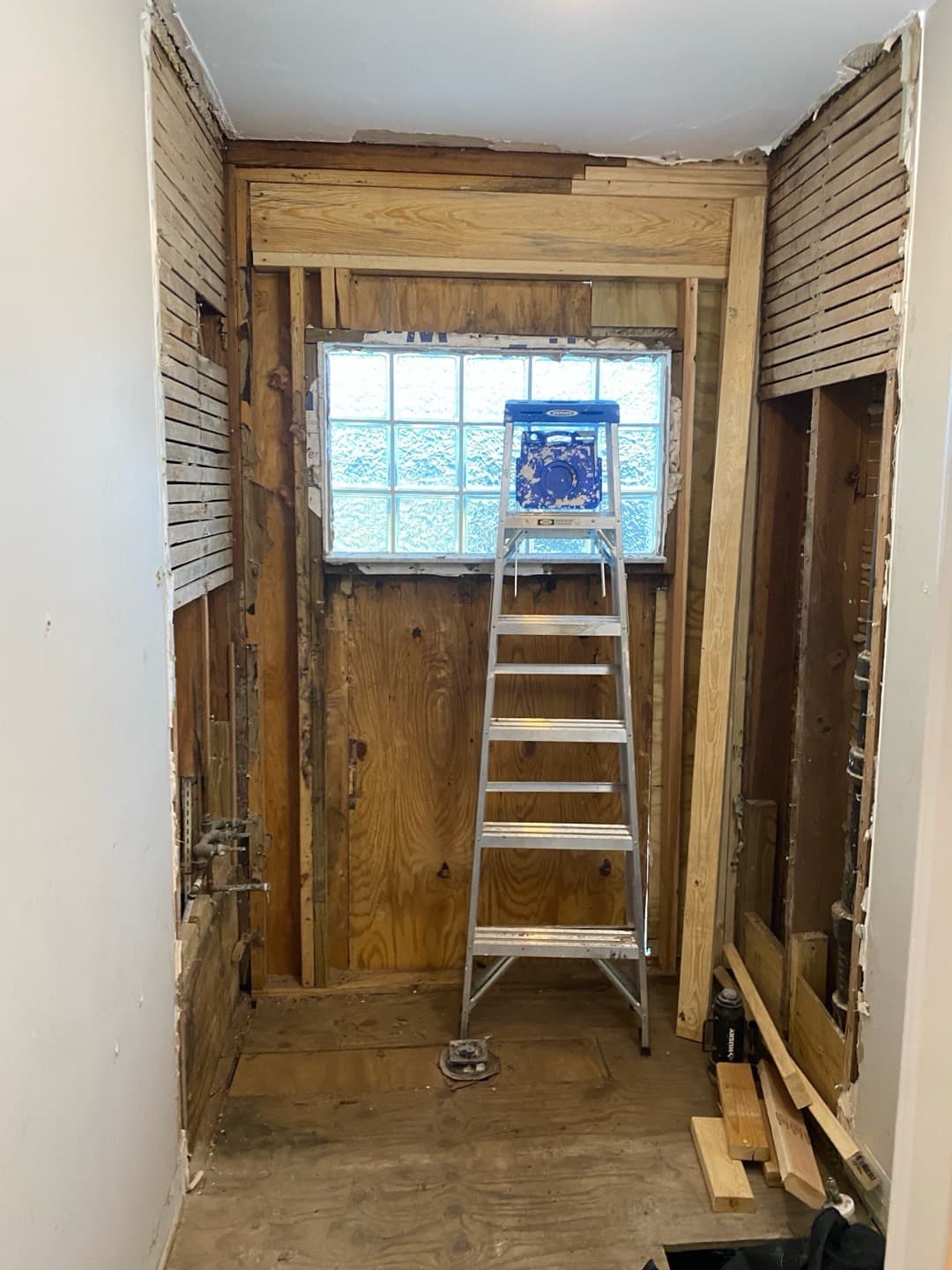 Wide demo view from the doorway showing the shower bay stripped to studs and subfloor, a stepladder in front of the glass-block window, and freshly sistered lumber framing the window opening (real Karhan project)